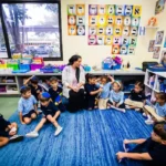 A teacher and students sitting in a classroom circle beneath Hebrew letter displays.