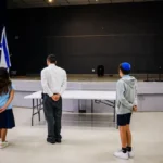 Students and a teacher facing the auditorium stage between Israeli and American flags.